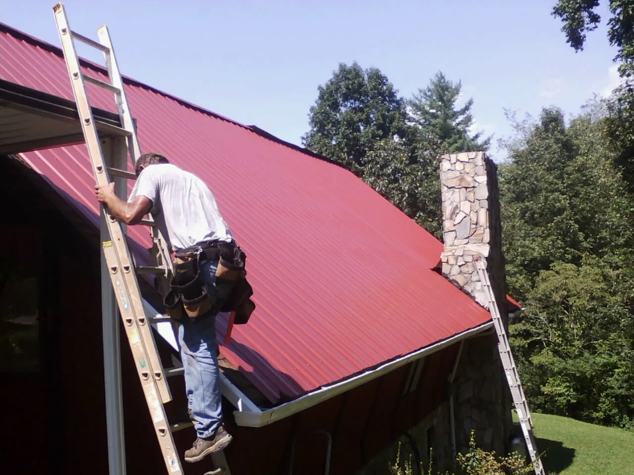 Roofer on a ladder beside a red metal roof and stone chimney — Soffit & Fascia Repair by Roofing Brothers in Center Line